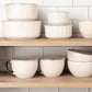 Set of ceramic bowls and mugs on a wooden shelf with a tiled wall background