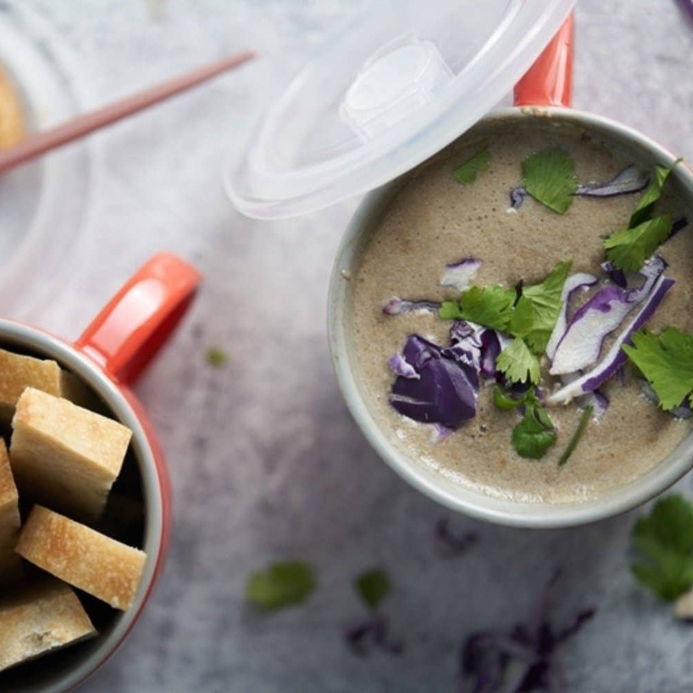 A styled overhead food scene showing a bowl of creamy mushroom soup with purple cabbage and cilantro garnish, alongside a bowl of bread cubes and a slice of bread on a light gray surface.