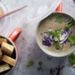 A styled overhead food scene showing a bowl of creamy mushroom soup with purple cabbage and cilantro garnish, alongside a bowl of bread cubes and a slice of bread on a light gray surface.