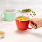 A red ceramic mug filled with yellow soup is held by a hand, with a translucent lid placed beside it. A mint green version of the mug and a small plate of green beans with red berries are visible in the background on a clean white surface.