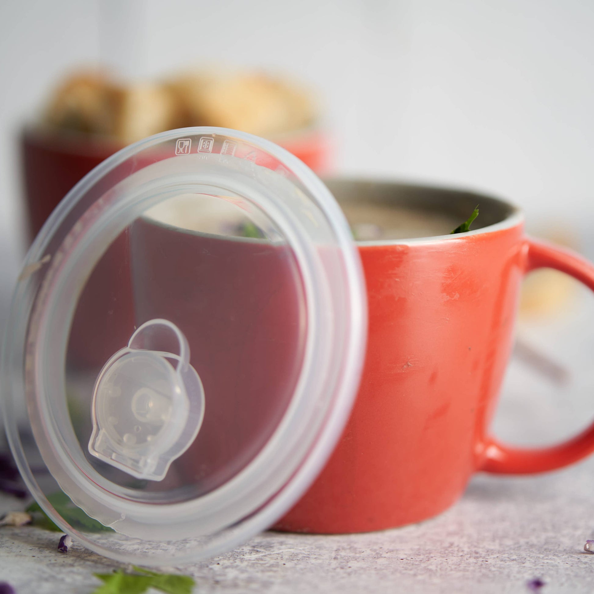 A clear silicone lid with a small clear plastic strainer or filter piece, shown leaning against an orange-red ceramic mug on a light tabletop.