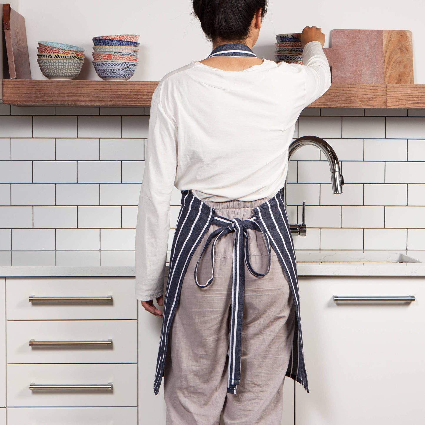 Person in a kitchen wearing an apron, standing by a shelf with bowls.