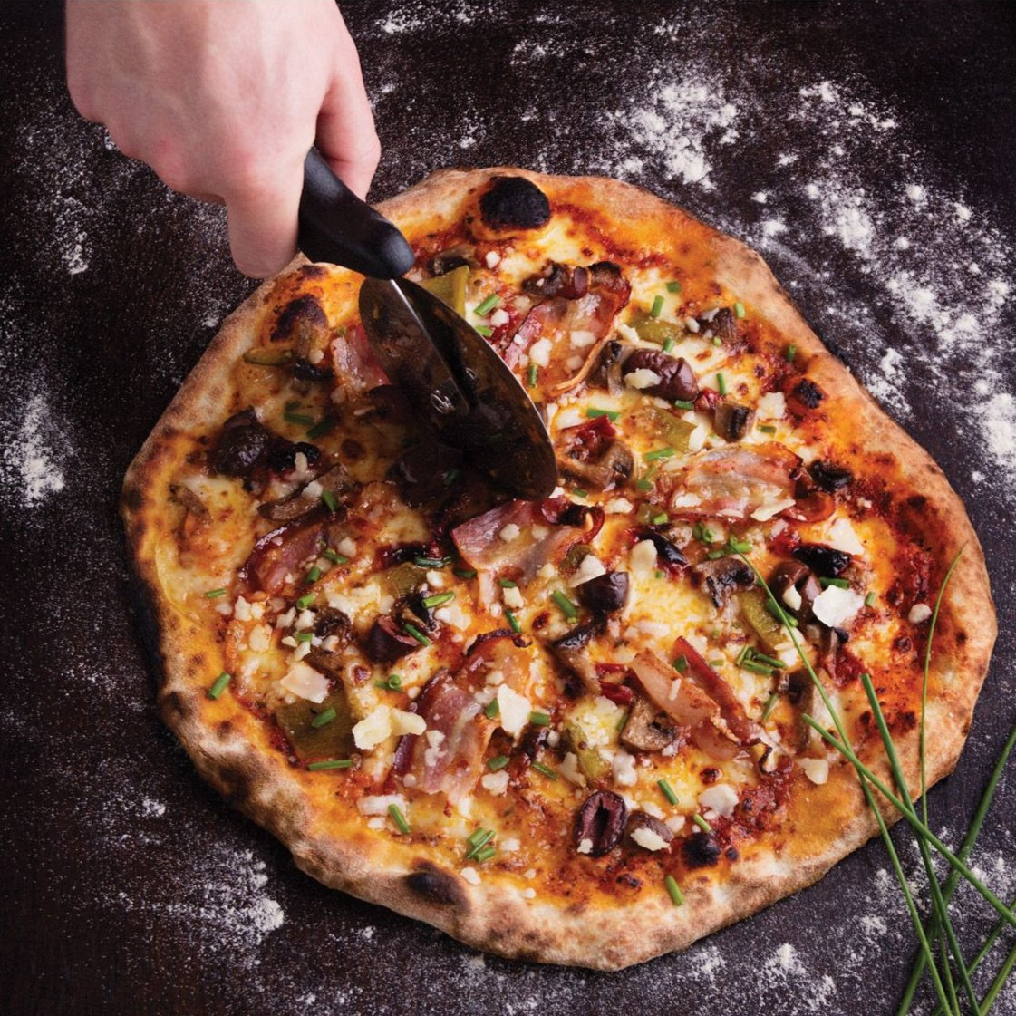 Classic Pizza Cutter being used to cut a pizza on a dark surface.