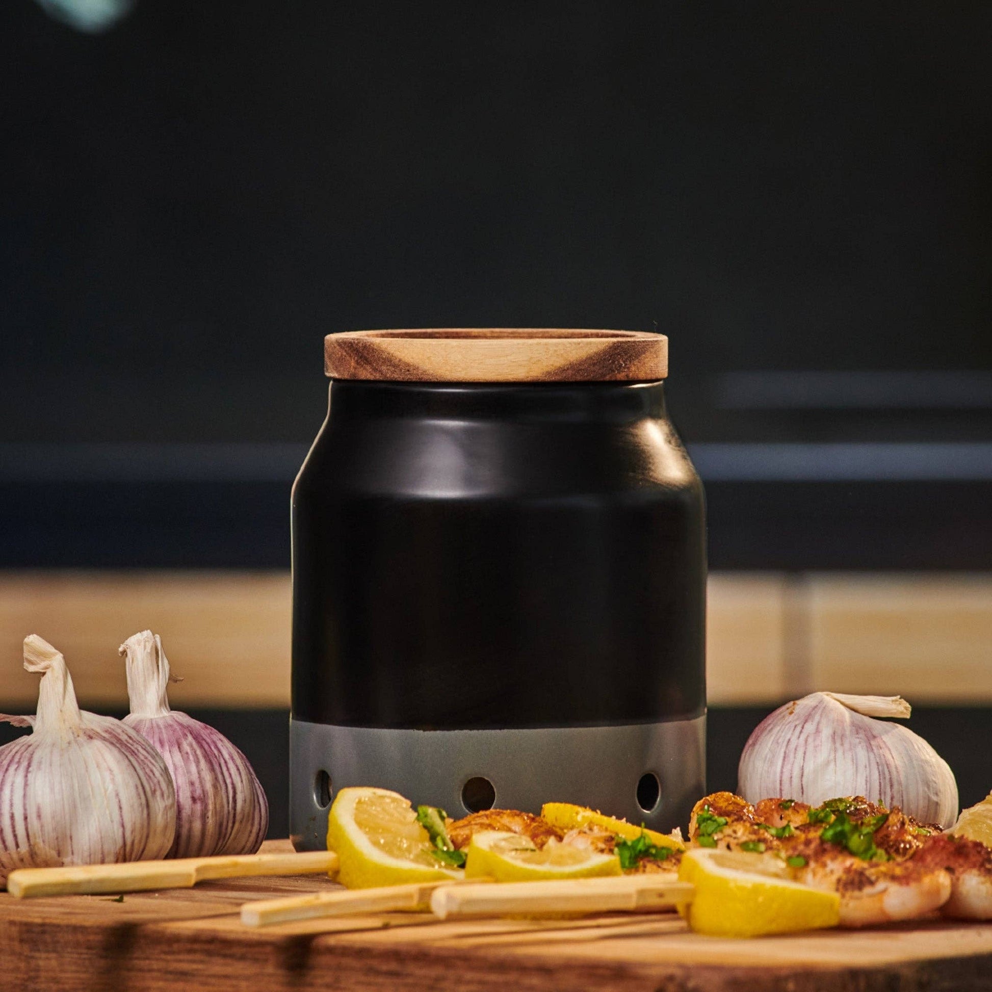 Black garlic keeper with wooden lid on a wooden surface with garlic and lemon slices.