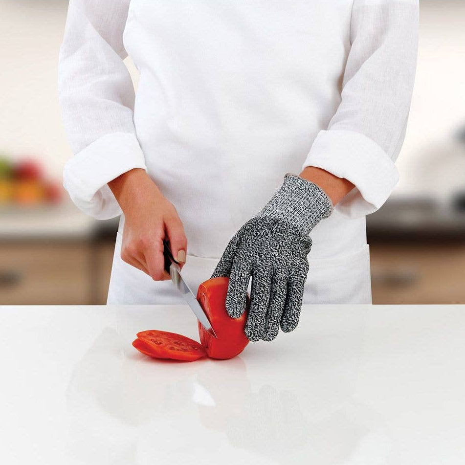 Person wearing a gray glove cutting a tomato on a white surface with a blurred kitchen background.