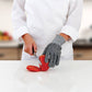 Person wearing a gray glove cutting a tomato on a white surface with a blurred kitchen background.