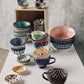 Collection of colorful ceramic bowls and mugs on a white surface with a concrete wall background.