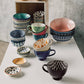 Collection of colorful ceramic bowls and mugs on a white surface with a concrete wall background.