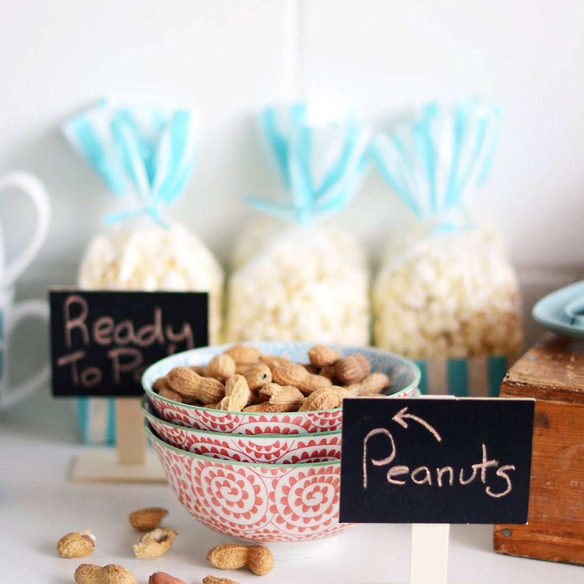 Bowl of peanuts with a 'Ready to Pop' sign and other snacks on a white surface.