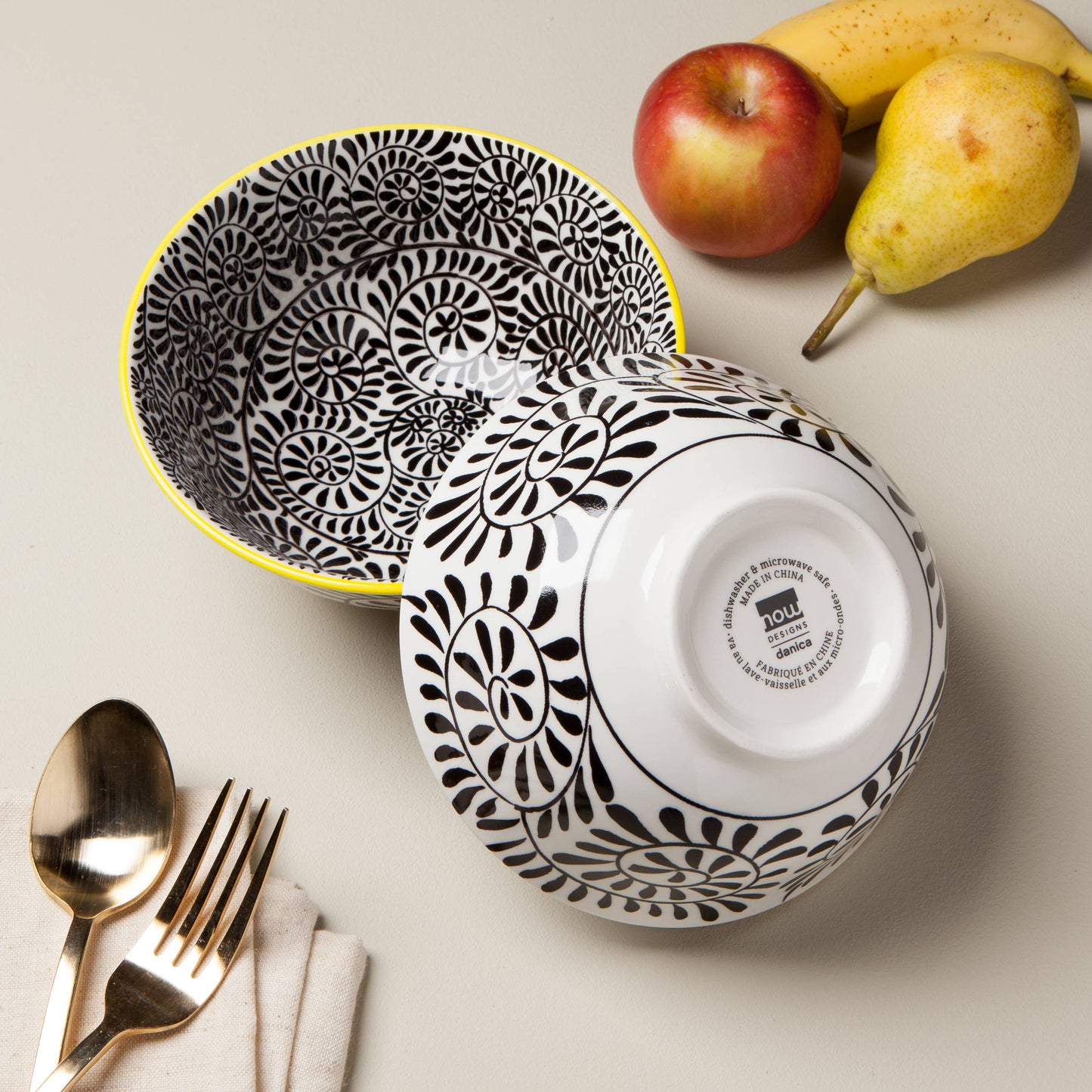 Black and white patterned bowls with a brand logo, apples, and bananas on a light surface.