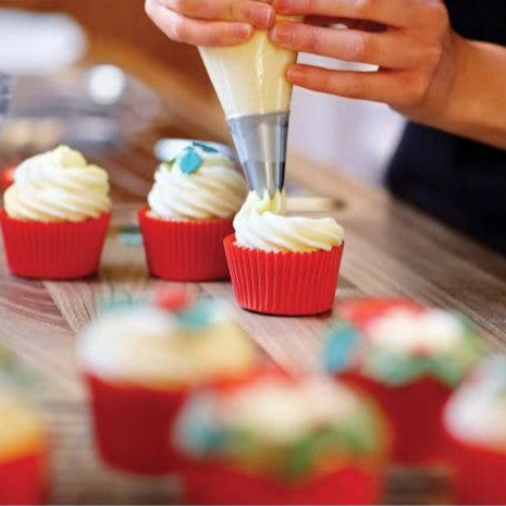 Person decorating cupcakes with white frosting using a piping bag on a wooden table.