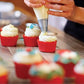 Person decorating cupcakes with white frosting using a piping bag on a wooden table.