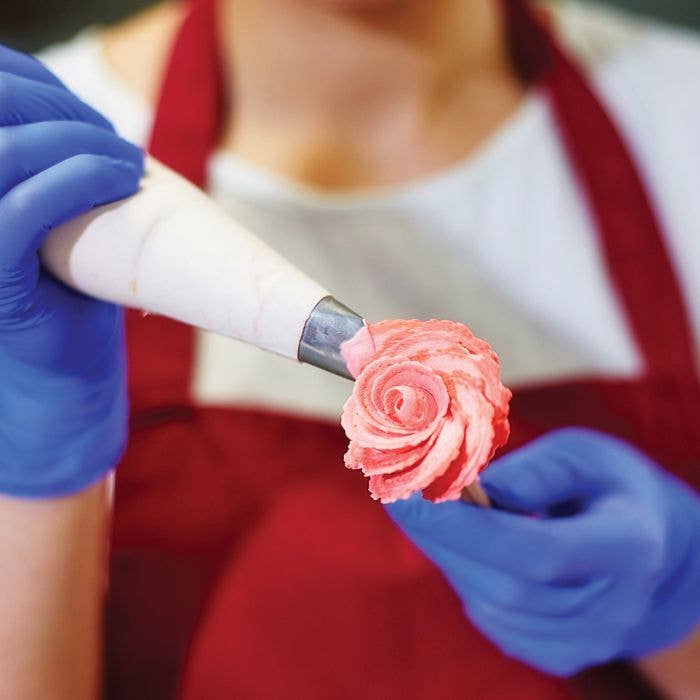 Person in blue gloves decorating a pink rose on a cake with a piping bag.