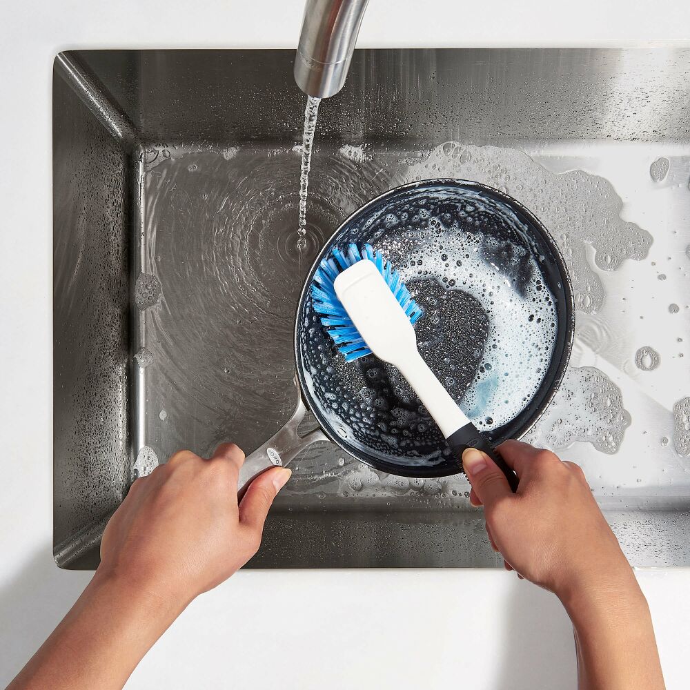 Hands washing a skillet in a metal sink with a white dish brush. 