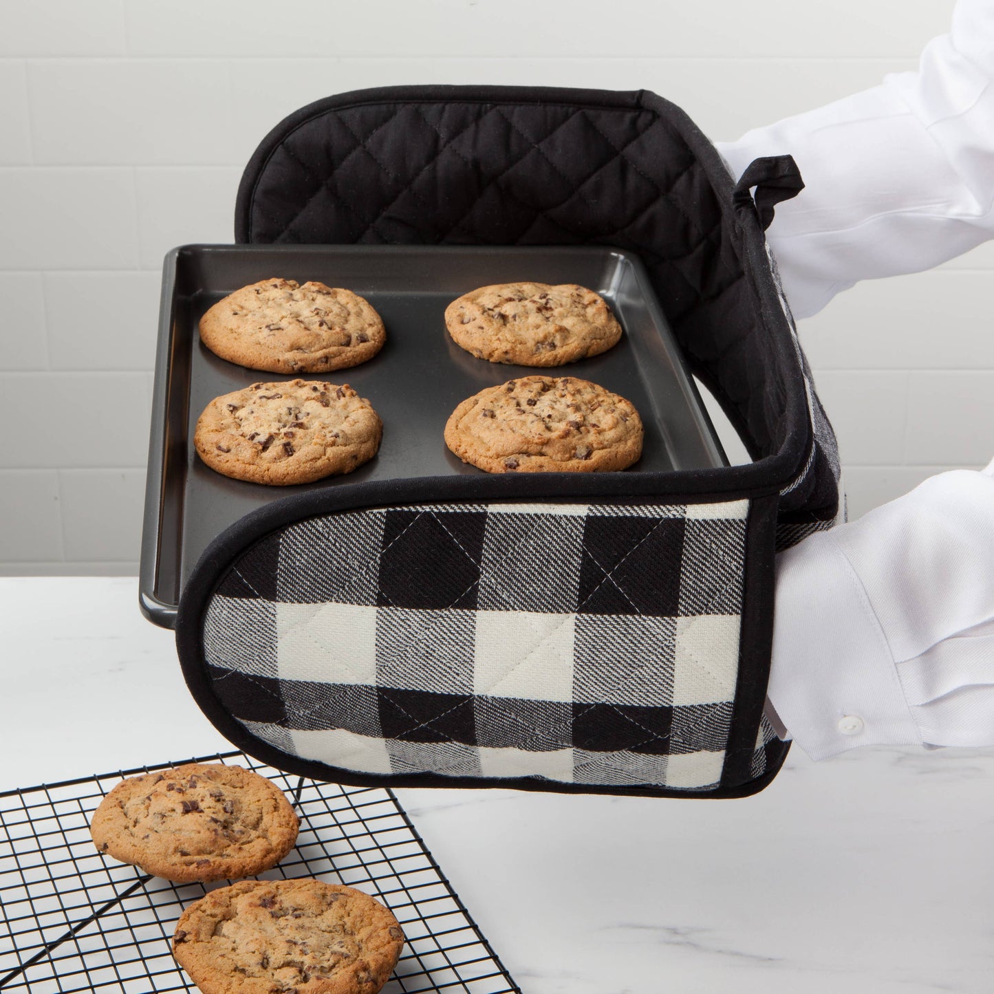 Black baking tray with cookies inside, held by a person wearing a white apron, on a light gray surface.