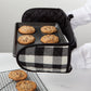 Black baking tray with cookies inside, held by a person wearing a white apron, on a light gray surface.