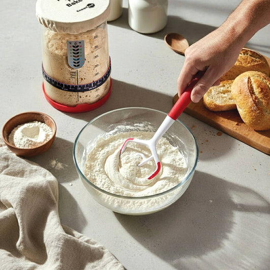 Person mixing ingredients in a bowl with a red whisk, surrounded by baking items on a light surface.