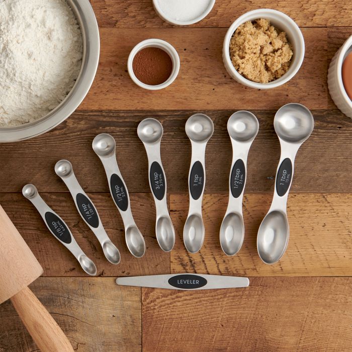 Set of measuring spoons on a wooden surface with ingredients in small containers.