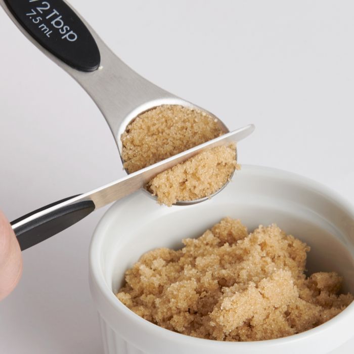 Measuring spoon filled with brown sugar being added to a white bowl.
