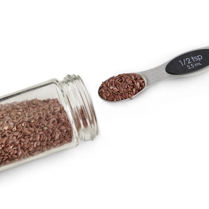 Glass jar filled with brown seeds and a spoon measuring 1/2 teaspoon next to it on a white background