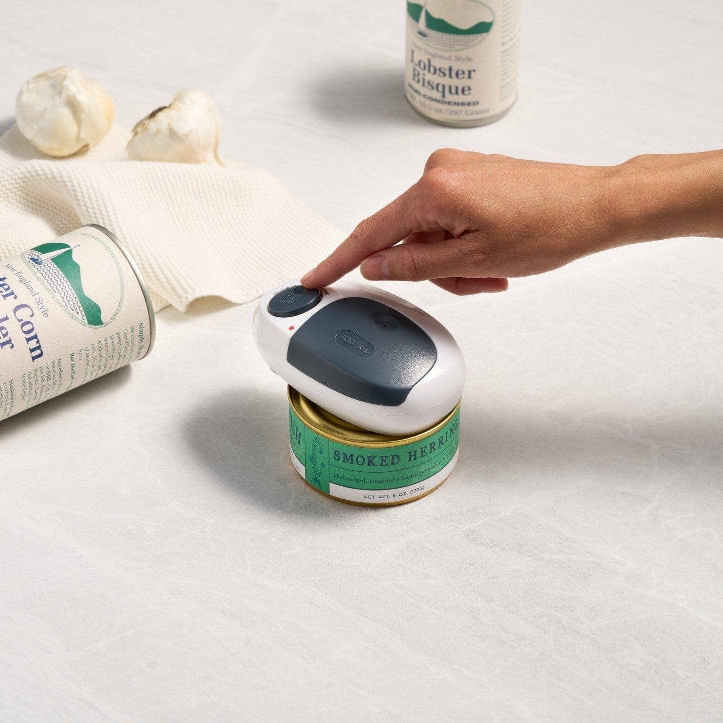 Hand interacting with a small electronic can opener on a white surface with cans in the background