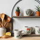 Set of mugs with floral designs on a wooden shelf with potted plants and wooden utensils.