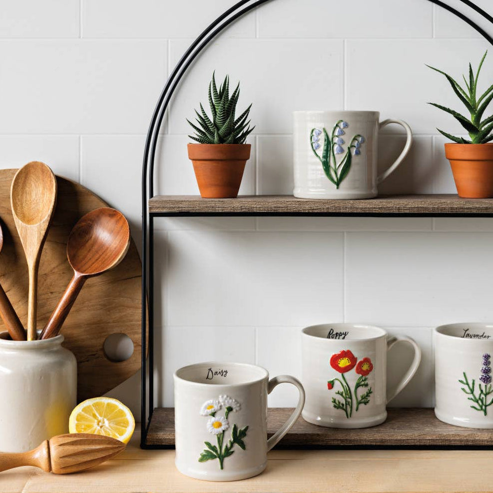 Set of mugs with floral designs on a wooden shelf with potted plants and wooden utensils.