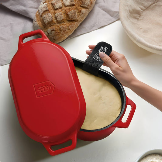 A red enameled cast iron Dutch oven with a lid, shown open with a hand holding a black oven mitt. Inside, a loaf of bread and dough are visible on a white countertop with a beige cloth in the background.