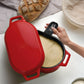 A red enameled cast iron Dutch oven with a lid, shown open with a hand holding a black oven mitt. Inside, a loaf of bread and dough are visible on a white countertop with a beige cloth in the background.