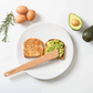 Avocado toast on a white plate with a wooden spreader, surrounded by avocados, eggs, and rosemary.