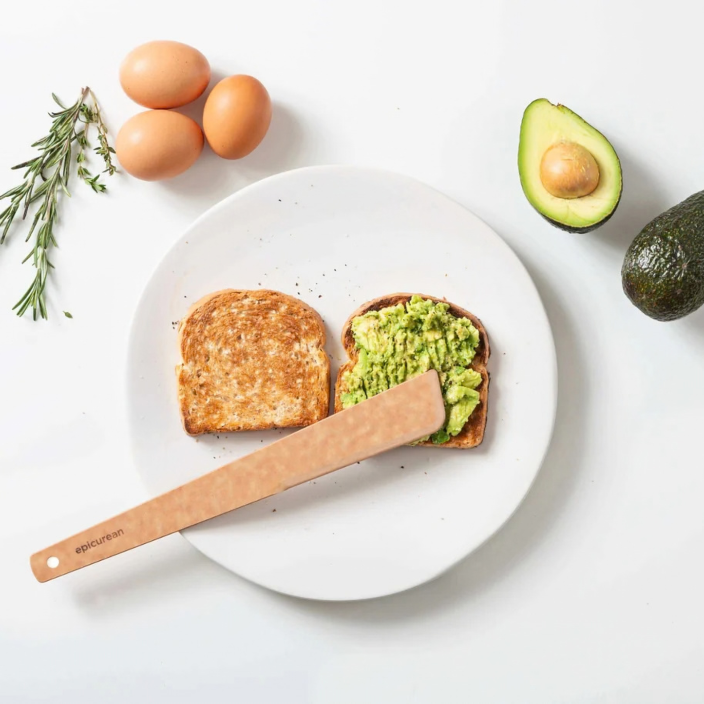 Avocado toast on a white plate with a wooden spreader, surrounded by avocados, eggs, and rosemary.
