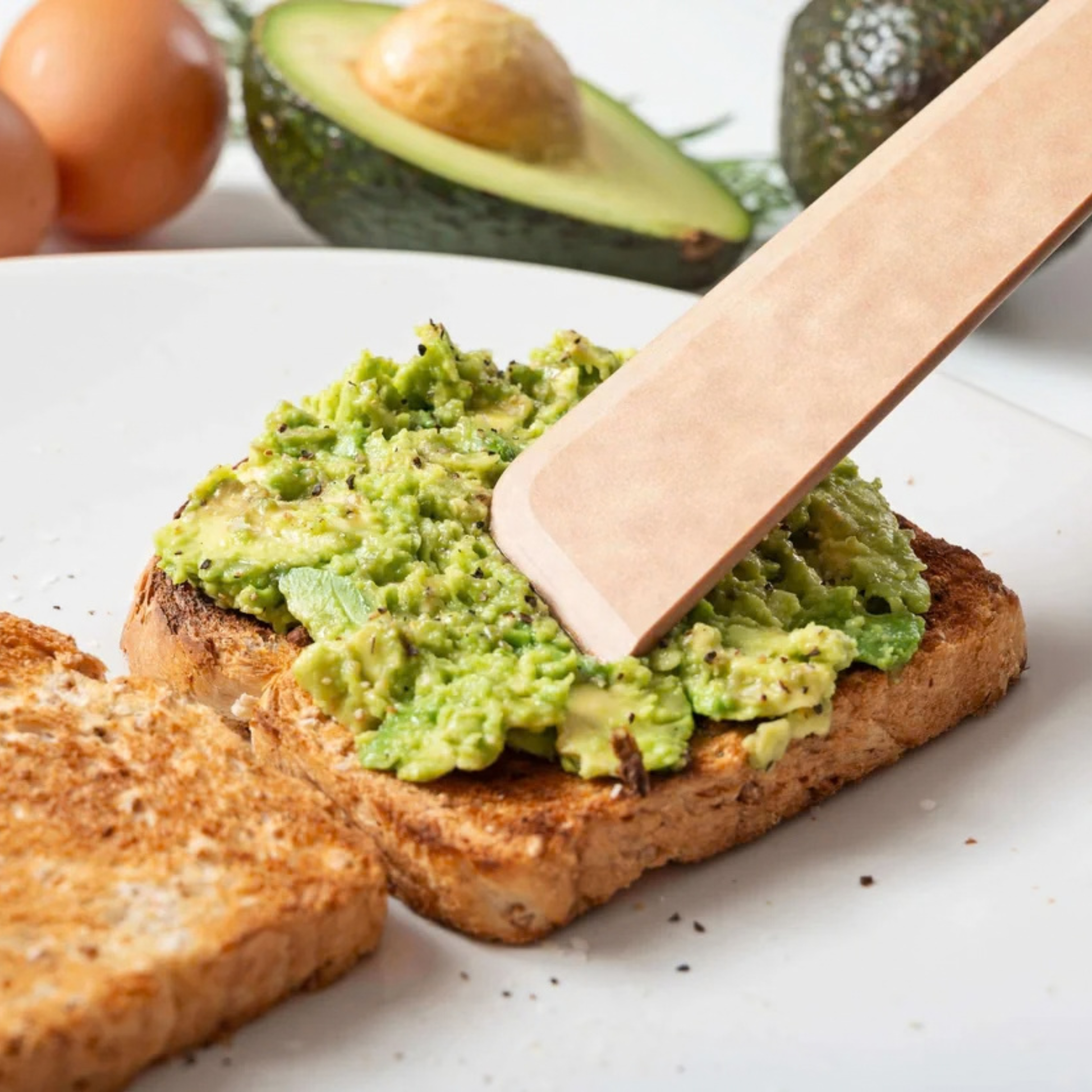 Avocado spread on toast with a wooden knife, surrounded by avocados and eggs.