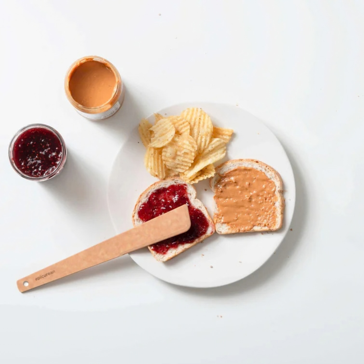 Peanut butter and jelly sandwich with chips on a white plate, surrounded by ingredients.