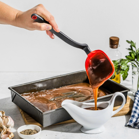 Hand using a ladle to pour brown liquid into a white gravy boat on a kitchen counter.