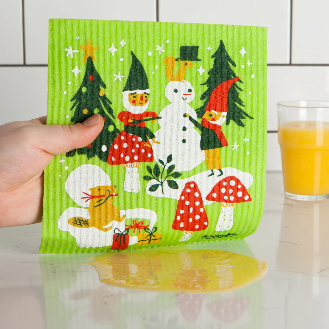 Green kitchen towel with Christmas-themed design held by a hand, with a glass of orange juice in the background.