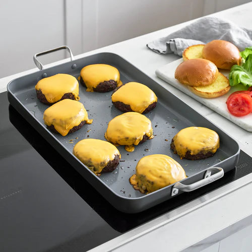Burgers with cheese on a griddle pan next to buns and vegetables on a kitchen counter.