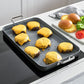 Burgers with cheese on a griddle pan next to buns and vegetables on a kitchen counter.