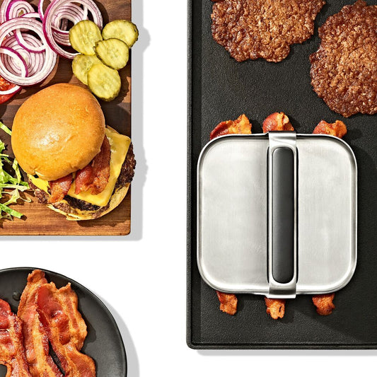 Top down view of a metal grill press being used to cook backon a a griddle. 
