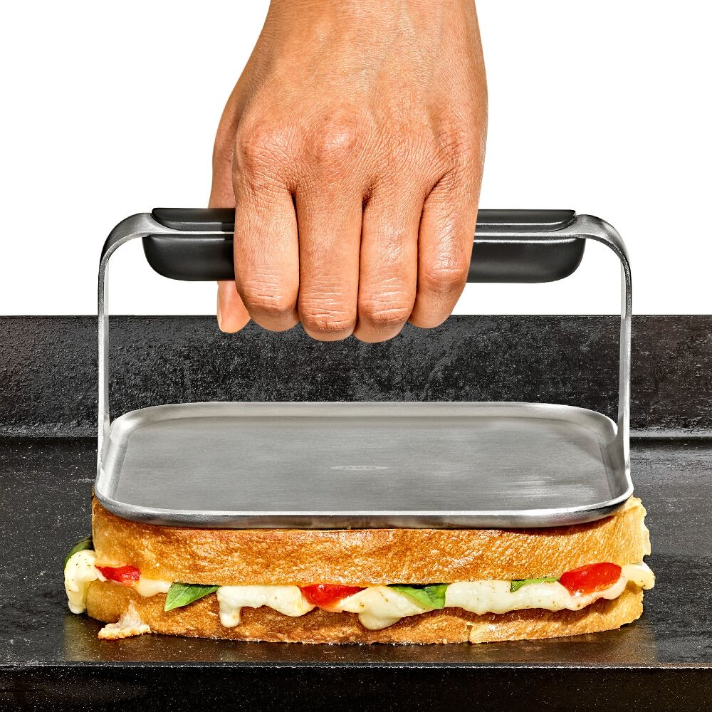 Hand using a griddle press on a cheesey sandwich on a black grill. 