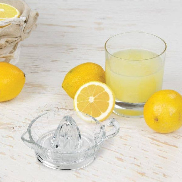 Clear glass lemon squeezer with lemons and a glass of lemon juice on a wooden surface.