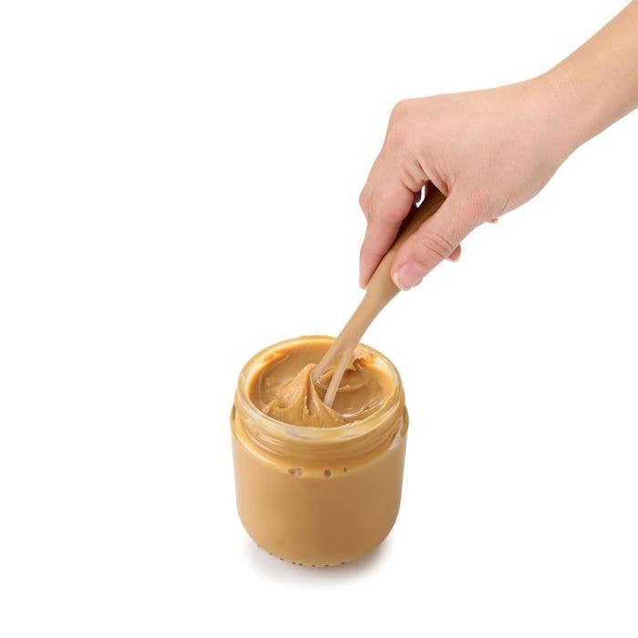 Hand using a wooden spoon to scoop peanut butter from a jar on a white background