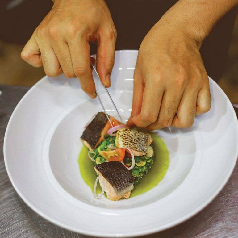 Person garnishing a dish with fish and vegetables on a white plate.