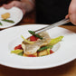 Person using a fork to eat a dish of fish and vegetables on a white plate.