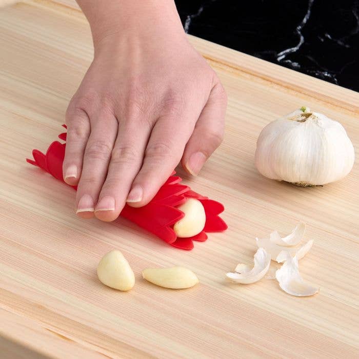 Person using a red garlic peeler on garlic cloves on a wooden cutting board.