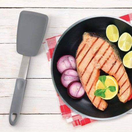 Frying pan with grilled salmon, lemon, and vegetables on a checkered cloth with a spatula.