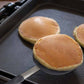 Three pancakes being cooked on a griddle with a spatula.