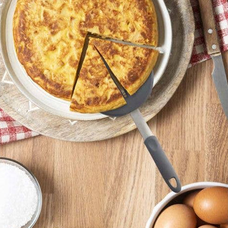 Omelette on a plate with a spatula, surrounded by potatoes and eggs on a wooden table.