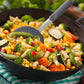 Frying pan with mixed vegetables and a spatula on a checkered tablecloth.