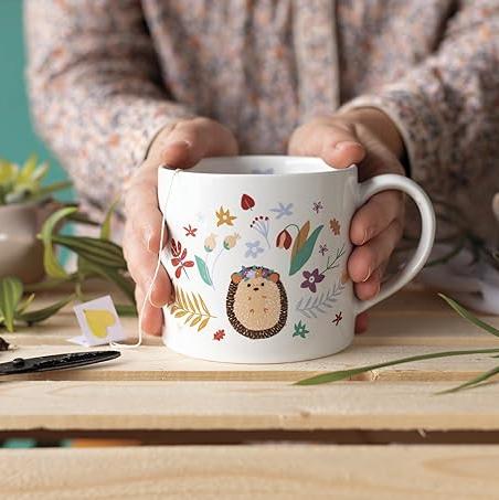  "Hedge Hugs" Mug in a Box brings a cozy, nature-inspired charm to your mornings. Featuring a whimsical hedgehog surrounded by wildflowers and a smooth white interior, 