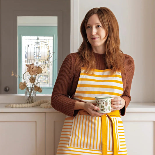 Woman wearing a yellow and white striped apron holding a mug in a kitchen.